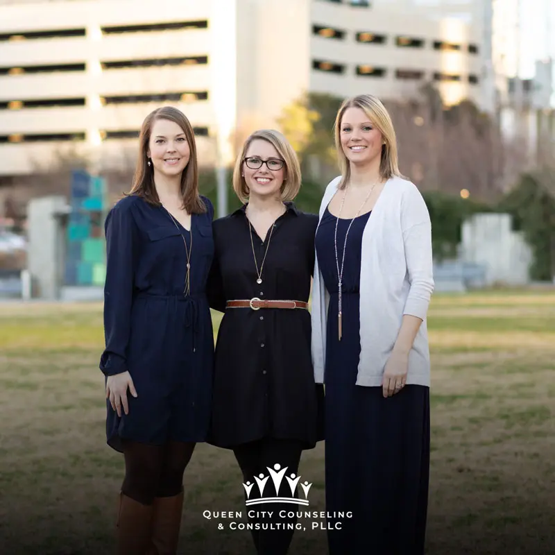 Three women standing side by side outdoors, smiling at the camera. They are dressed in dark dresses, and one wears a light cardigan. Buildings and trees are visible in the background. Queen City Counseling & Consulting, PLLC is at the bottom.